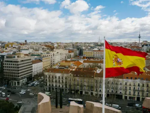 A Spanish flag waves in the wind against a cityscape