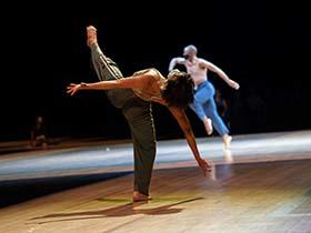 A dancer, who is facing away from the camera, is bending backwards with arms outstretched, and their left leg raised high above their waist. Another dancer is moving in the background. Both are wearing masks.
