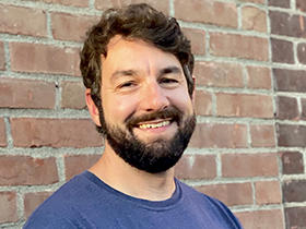 Hugh Martin, wearing a blue t-shirt, smiles and stands in front of a brick wall.