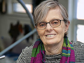 Susan Schmidt, wearing a black and white shirt and a colorful scarf, smiles while holding onto a banister.