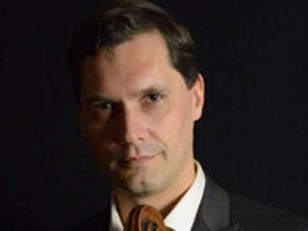 Jan Muller-Szeraws, wearing a suit, smiles into the camera with part of his cello visible.