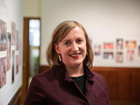Meredith Fluke, wearing a maroon jacket and smiling into the camera, stands in a room with pictures on the walls that have been blurred by the camera.