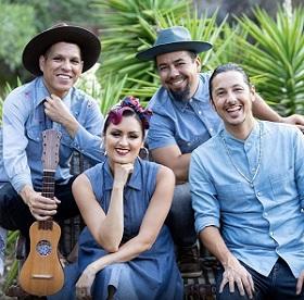Three men and a woman, all in denim, sitting outside on a bench smiling and looking into the camera. One holds a stringed instrument