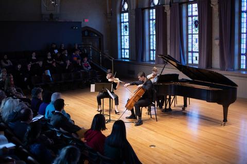 Photo of three musicians, performing in Brooks Concert Hall on the piano, cello and violin with the audience watching