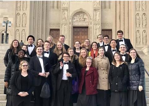 A group photo of college choir with Allegra Martin, choir director, all smiling with the backdrop of the front entrance to St. Joseph Chapel