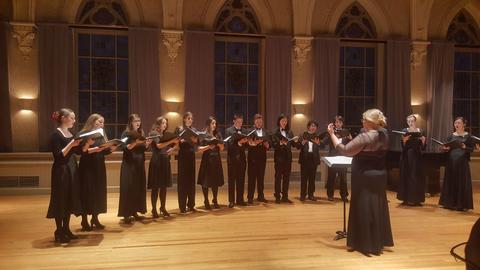 Photo taken from a concert performance with 14 members of the ensemble standing in a semi circle with choir director Allegra Martin conducting.  All members are dressed in black choir attire
