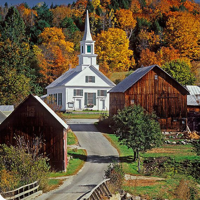 A picturesque image of a New England town in the fall. There is a one-lane road with wooden barns on either side and a white church at the end of the road. Orange and yellow trees with fall foliage are in the background