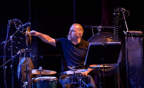 An action photo of Stuart Gerber playing several percussion instruments and drums, wearing a black t-shi
