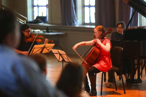  Photo of three students in a recital, seated, playing cello, violin and piano