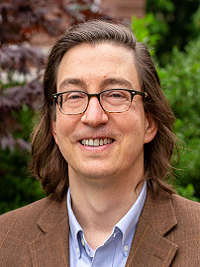 faculty headshot man with medium color hair and length with glasses and brown suit coat blue shirt and green leaves in back of him