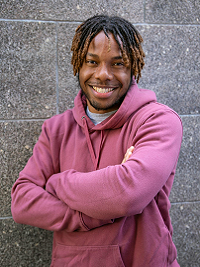 close up of a man with his arms crossed in front braids in his hair wearing a light red shirt standing in front of a wall