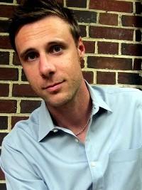 close up of man with dark hair light blue button down shirt with a brick wall as the background