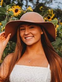 close up of a girl wearing a floppy hat white dress long redish brown hair smiling.  There are sunflowers behind her.