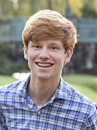young man with light red hair wearing a blue and white plaid shirt smiling at the camera with a building in the background
