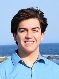 young man standing on a beach wearing a light blue shirt and short brown hair smiling at the camera