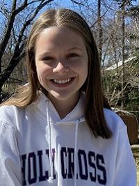 young woman wearing a white sweatshirt that says Holy Cross, she has medium length brown hair and is smiling