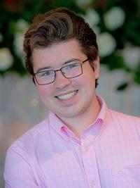young man wearing glasses and a pink button down shirt he has short brown hair and smiling at the camera