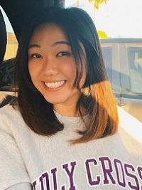 young woman with medium length hair brown in color sitting wearing a gray Holy Cross sweatshirt smiling at the camera