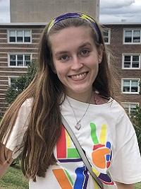 young woman in front of a brick building wearing a white tshirt with bright colors on it smiling at the camera