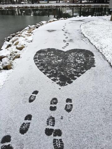 A snowy scene by a pond, white pathway lightly dusted with snow, a black heart shape where footsteps have stamped the path free of snow.  Black footsteps lead to and from the heart.
