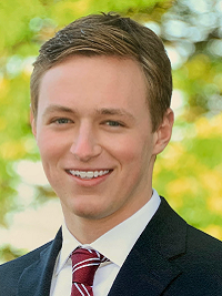 close up of a young man outside wearing a tie and dark blazer smiling at the camera 