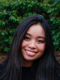 close up of a young woman standing outside, she has long black hair and is smiling at the camera