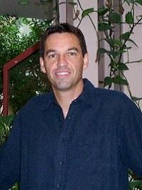man smiling at the camera wearing a dark blue shirt with plants behind him