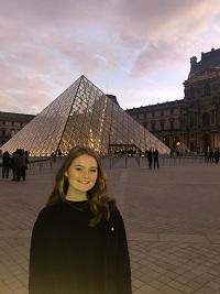 Lorna, in front of the glass pyramid at the Louvre Museum, smiles at the camera. Lorna has light colored hair and is wearing dark clothes. 