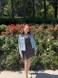 Allison stands in front of blooming rose bushes and smiles at the camera. Alison has short dark hair and wears a jean jacket and dress. 