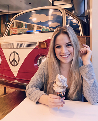 Chloe, who has blond hair and wears a grey shirt, sits in front of an old VW van that is inside a building. She smiles holding an ice cream cone. 