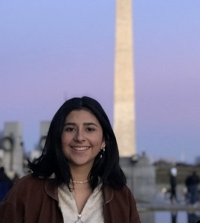 Standing in front of an outdoor tourist site, Daniela smiles. Daniela has dark hair and is wearing a brown coat over a white shirt.