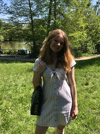 Standing in front of trees and a pond, Lorna smiles. Lorna has strawberry blond hair and wears a striped white and black dress and holds a bag. 