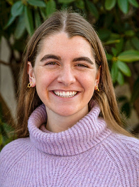In this headshot, Maggie, who has brown hair and is wearing a purple turtleneck sweater, looks at the camera and smiles. 