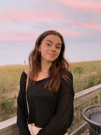 Michaela wears a black shirt, has brown hair, and smiles at the camera. It is sunset, and Michaela stands outside in front of some grass and clouds catching the sun.