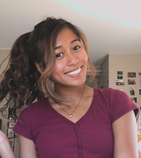 Serey wears a maroon t-shirt, and her dark curly hair is visible in a flowing ponytail. Serey smiles at the camera and behind her is a wall that has photos on it.