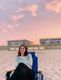 Michaela is sitting in a chair on the beach during sunset and smiling. She has brown hair and is wearing a green shirt and black pants.