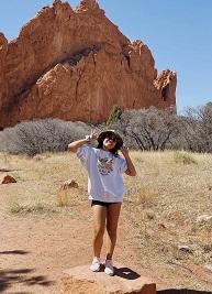 Serey is standing on a rock in front of a natural rock formation. She wears a hat and sunglasses. 