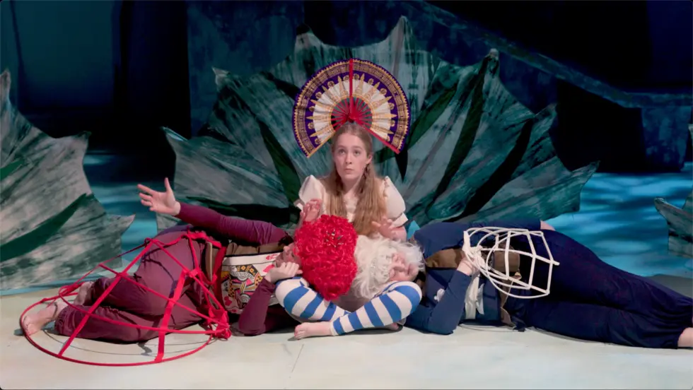 Blond woman sitting on stage, while two other people lay on the stage