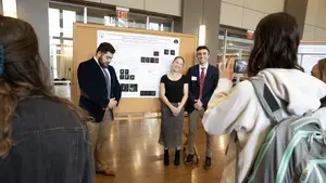 three students having their photo taken in front of a presentation board during the academic conference