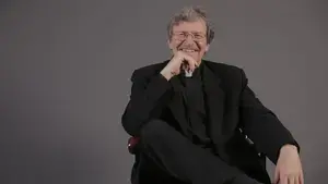 Photo of William A. Clark, S.J. sitting and smiling with one leg crossed and a hand on his chin.